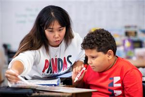 Female, teacher with long dark hair working with a male 4th grade student at his desk.